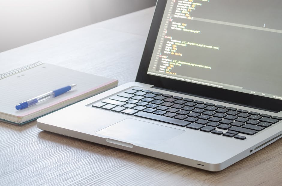 A close-up view of a silver laptop open on a light wooden desk, displaying lines of code on its screen, next to a spiral-bound notebook with a blue pen resting on top. The background is softly lit, em