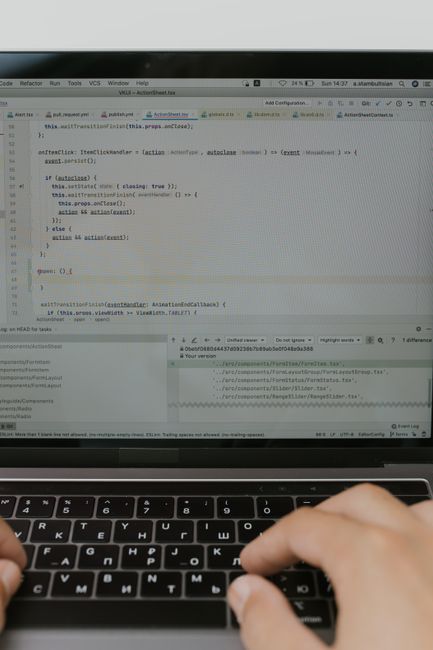 A close-up view of a silver laptop open on a light wooden desk, displaying lines of code on its screen, next to a spiral-bound notebook with a blue pen resting on top. The background is softly lit, em